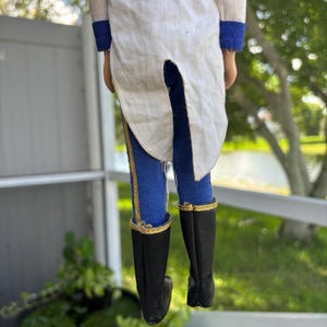 May include: A vintage doll wearing a white jacket with gold trim and blue pants with gold stripes. The doll has black boots with gold trim. The doll is standing in front of a white fence and green foliage.