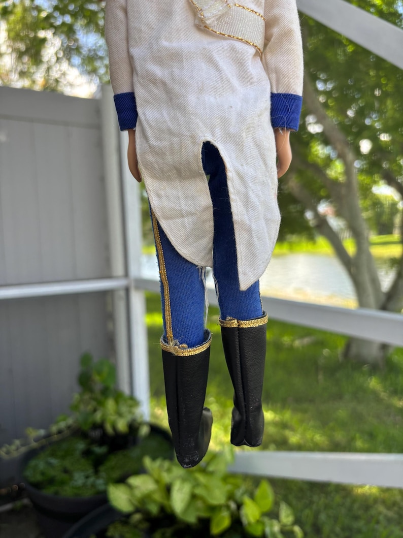 May include: A vintage doll wearing a white jacket with gold trim and blue pants with gold stripes. The doll has black boots with gold trim. The doll is standing in front of a white fence and green foliage.