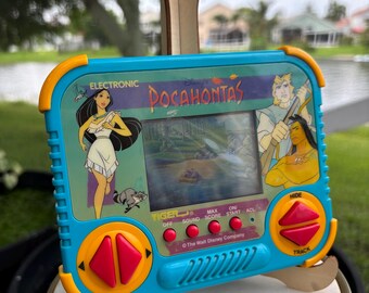 Op de afbeelding: Een vintage elektronisch handheld spel van Tiger Electronics met Disney's Pocahontas. Het apparaat is lichtblauw met gele accenten, roze knoppen en een scherm dat een spelscène toont. Het bevat illustraties van Pocahontas, John Smith en Kocoum.