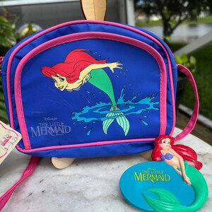 Op de afbeelding: Een blauwe schoudertas met Ariel uit Disney's De Kleine Zeemeermin, met roze rand en riem. Er is ook een bijpassend Ariel-beeldje aanwezig. De tas heeft een houten handvat.