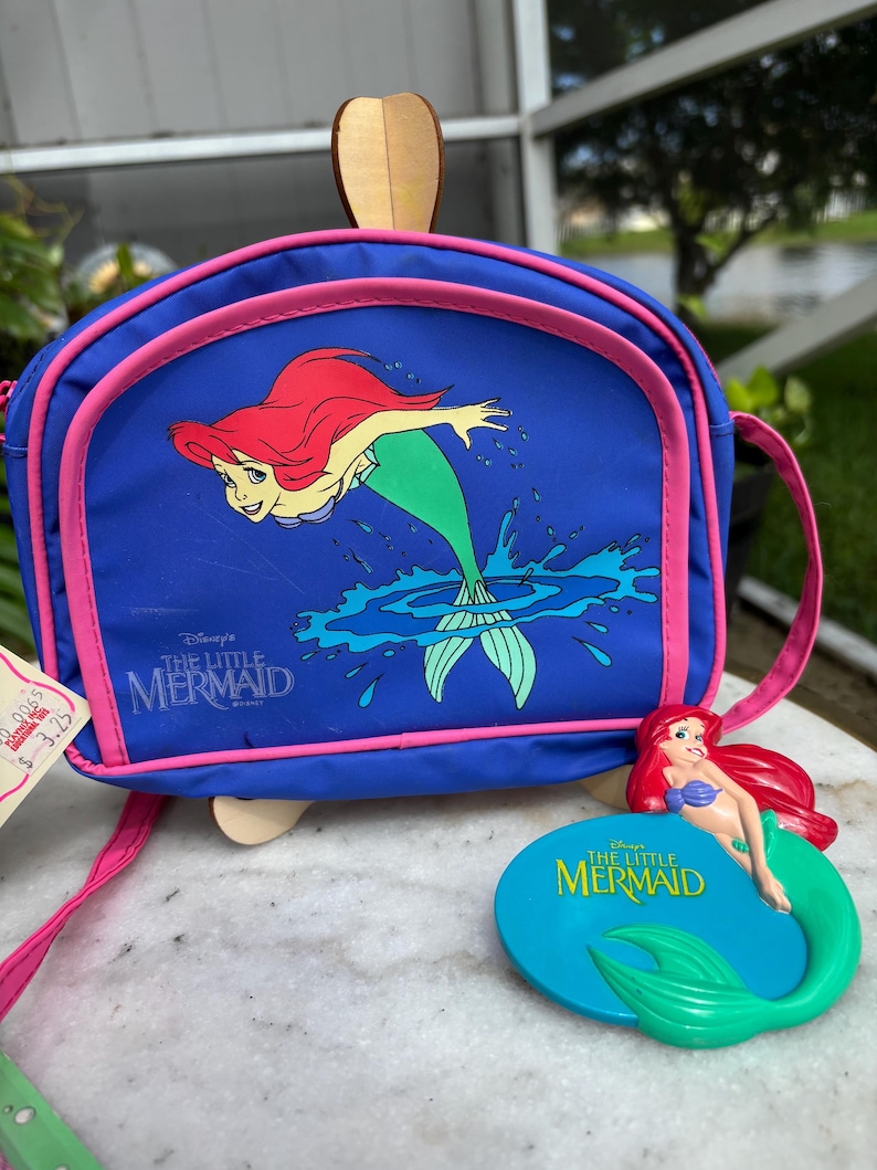 Op de afbeelding: Een blauwe schoudertas met Ariel uit Disney's De Kleine Zeemeermin, met roze rand en riem. Er is ook een bijpassend Ariel-beeldje aanwezig. De tas heeft een houten handvat.