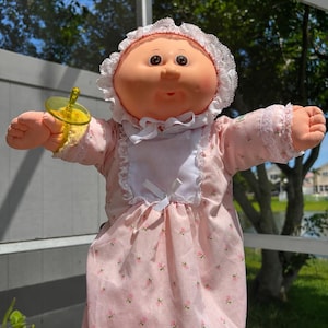 Pode incluir: Uma boneca com rosto redondo cor de pêssego, olhos escuros e bochechas rosadas, vestindo um vestido rosa com estampa floral pequena, um babador branco com laço e uma touca branca com renda. A boneca segura uma chupeta de plástico amarela.
