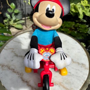 May include: A Mickey Mouse toy figure riding a red tricycle. The figure has a red hat, black ears, and white gloves. The tricycle has a black front tire and yellow footrests. The toy is set against a blurred background.