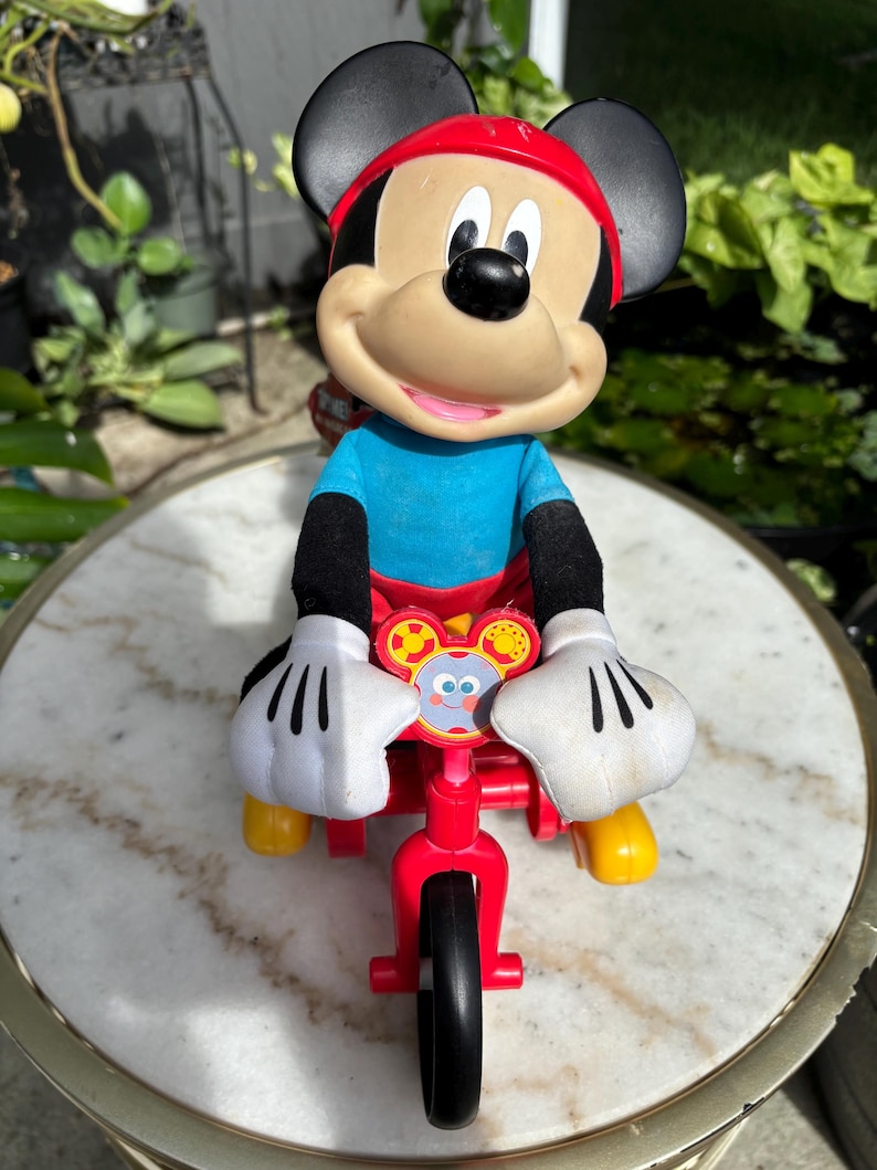 May include: A Mickey Mouse toy figure riding a red tricycle. The figure has a red hat, black ears, and white gloves. The tricycle has a black front tire and yellow footrests. The toy is set against a blurred background.