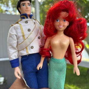 May include: Two dolls, a prince in a white jacket with gold trim and blue pants, and a mermaid with red hair and a green tail. The dolls are held in front of a blurred green background.