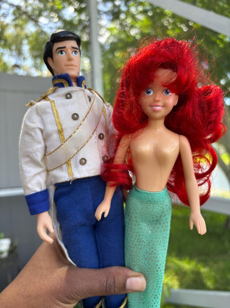 May include: Two dolls, a prince in a white jacket with gold trim and blue pants, and a mermaid with red hair and a green tail. The dolls are held in front of a blurred green background.