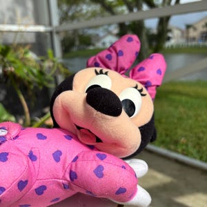 Pu&ograve; includere: Un pupazzo di Minnie Mouse in posizione sdraiata. La bambola ha una testa nera, un fiocco rosa con cuori viola e un corpo rosa con motivi a cuore viola. Il viso &egrave; rosa chiaro con grandi occhi e un naso nero.