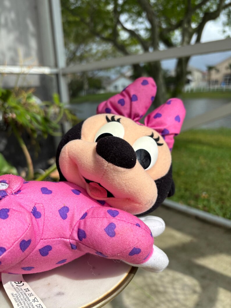 Pu&ograve; includere: Un pupazzo di Minnie Mouse in posizione sdraiata. La bambola ha una testa nera, un fiocco rosa con cuori viola e un corpo rosa con motivi a cuore viola. Il viso &egrave; rosa chiaro con grandi occhi e un naso nero.