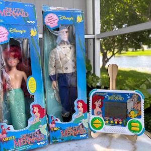 May include: Two vintage Disney's The Little Mermaid dolls in their original boxes, Ariel and Eric, along with an electronic handheld game. The Ariel doll has red hair and a green tail. The Eric doll is wearing a cream jacket and blue pants. The game features Ariel and other characters.