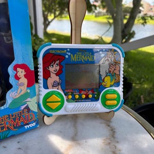 May include: A vintage electronic handheld game featuring Ariel from Disney's The Little Mermaid. The game has a white and blue casing with character graphics and button controls. The game is displayed on a wooden stand.