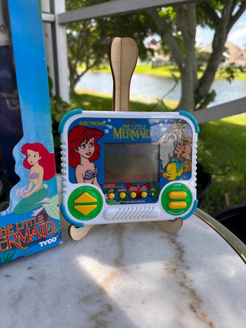 May include: A vintage electronic handheld game featuring Ariel from Disney's The Little Mermaid. The game has a white and blue casing with character graphics and button controls. The game is displayed on a wooden stand.