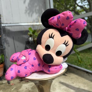Pu&ograve; includere: Una bambola di peluche di Minnie Mouse in posa reclinata. La bambola indossa un completo rosa con motivi a cuore viola e un fiocco abbinato. La bambola ha orecchie nere, naso nero e grandi occhi bianchi.