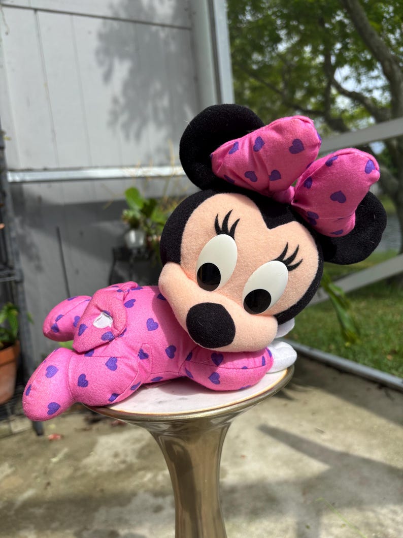 Pu&ograve; includere: Una bambola di peluche di Minnie Mouse in posa reclinata. La bambola indossa un completo rosa con motivi a cuore viola e un fiocco abbinato. La bambola ha orecchie nere, naso nero e grandi occhi bianchi.