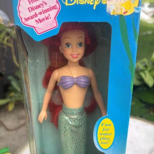 Pode incluir: Uma boneca Ariel da Disney com cabelos longos vermelhos, um top de conchas roxo e uma cauda de sereia verde cintilante, embalada numa caixa azul com o texto From Disney's Award-winning Movie! e Fun for water play, too! e uma etiqueta amarela.