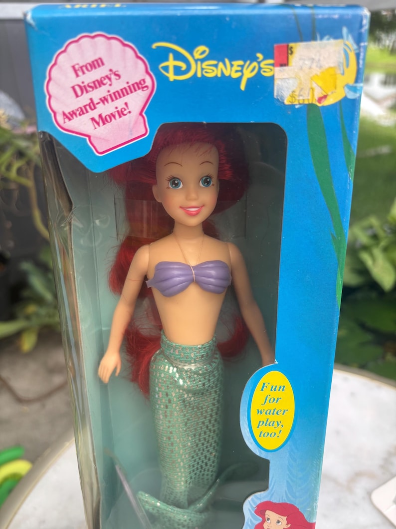 Pode incluir: Uma boneca Ariel da Disney com cabelos longos vermelhos, um top de conchas roxo e uma cauda de sereia verde cintilante, embalada numa caixa azul com o texto From Disney's Award-winning Movie! e Fun for water play, too! e uma etiqueta amarela.