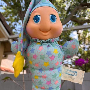 May include: A vintage Playskool Glo Worm plush toy. The toy has a light blue body and cap, adorned with colorful stars. It features a smiling face with large blue eyes and a yellow star on the end of its cap. A tag is attached.
