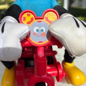 May include: A red tricycle toy with a Mickey Mouse figure. The toy features a red frame, black wheels, and yellow footrests. The Mickey Mouse figure wears a blue shirt, black gloves, and yellow shoes. The tricycle has a decorative face with a smiling face.