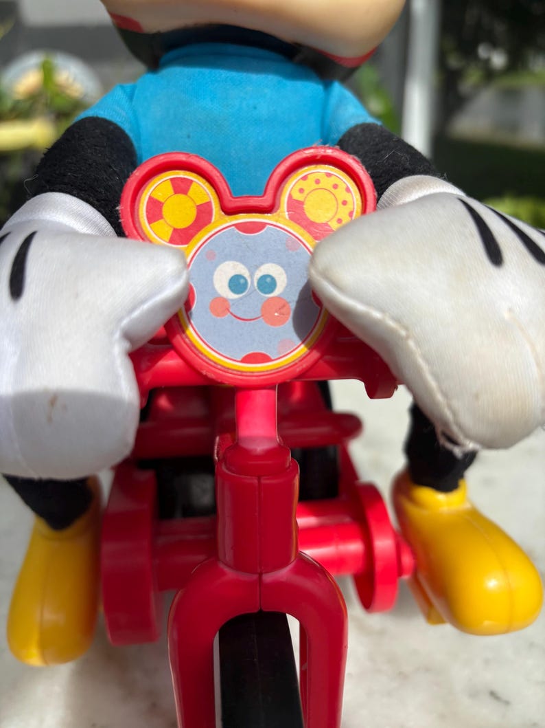 May include: A red tricycle toy with a Mickey Mouse figure. The toy features a red frame, black wheels, and yellow footrests. The Mickey Mouse figure wears a blue shirt, black gloves, and yellow shoes. The tricycle has a decorative face with a smiling face.