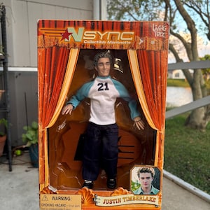 Może przedstawiać: Kolekcjonerska marionetka Nsync Justina Timberlake'a w oryginalnym opakowaniu. Pudełko ma wzór kurtyny scenicznej z logo Nsync. Lalka ma na sobie biało-niebieską koszulę z numerem 21, ciemne spodnie i czarne buty.