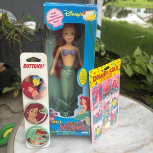 Pode incluir: Uma boneca Ariel da Pequena Sereia da Disney numa caixa azul, um cart&atilde;o vertical com tr&ecirc;s bot&otilde;es circulares com a Ariel e o Linguado, e uma folha de autocolantes Disney Star com v&aacute;rias ilustra&ccedil;&otilde;es de personagens, tudo na embalagem original.