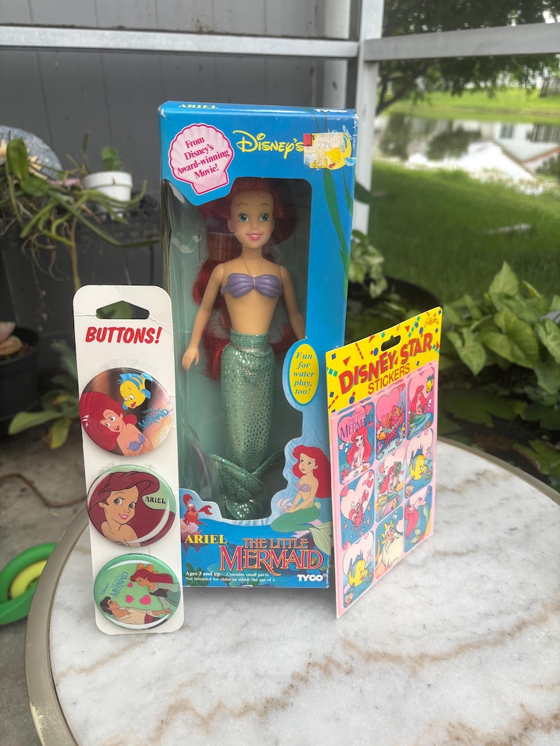 Pode incluir: Uma boneca Ariel da Pequena Sereia da Disney numa caixa azul, um cart&atilde;o vertical com tr&ecirc;s bot&otilde;es circulares com a Ariel e o Linguado, e uma folha de autocolantes Disney Star com v&aacute;rias ilustra&ccedil;&otilde;es de personagens, tudo na embalagem original.