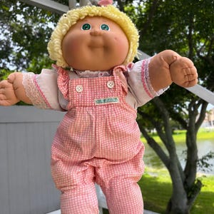 Könnte beinhalten: Eine Puppe mit rundem Gesicht, Grübchen an den Wangen und grünen Augen, mit lockigem gelbem Wollhaar, das mit einem kleinen grauen und rosafarbenen Accessoire verziert ist. Sie trägt ein weißes Hemd mit Spitzenbesatz und eine rosa-weiß karierte Latzhose.