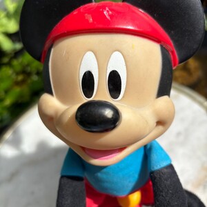 May include: A Mickey Mouse figurine with a red cap, black ears, and a blue shirt. The toy is riding a red and yellow bicycle. The figure has a black nose and white eyes.