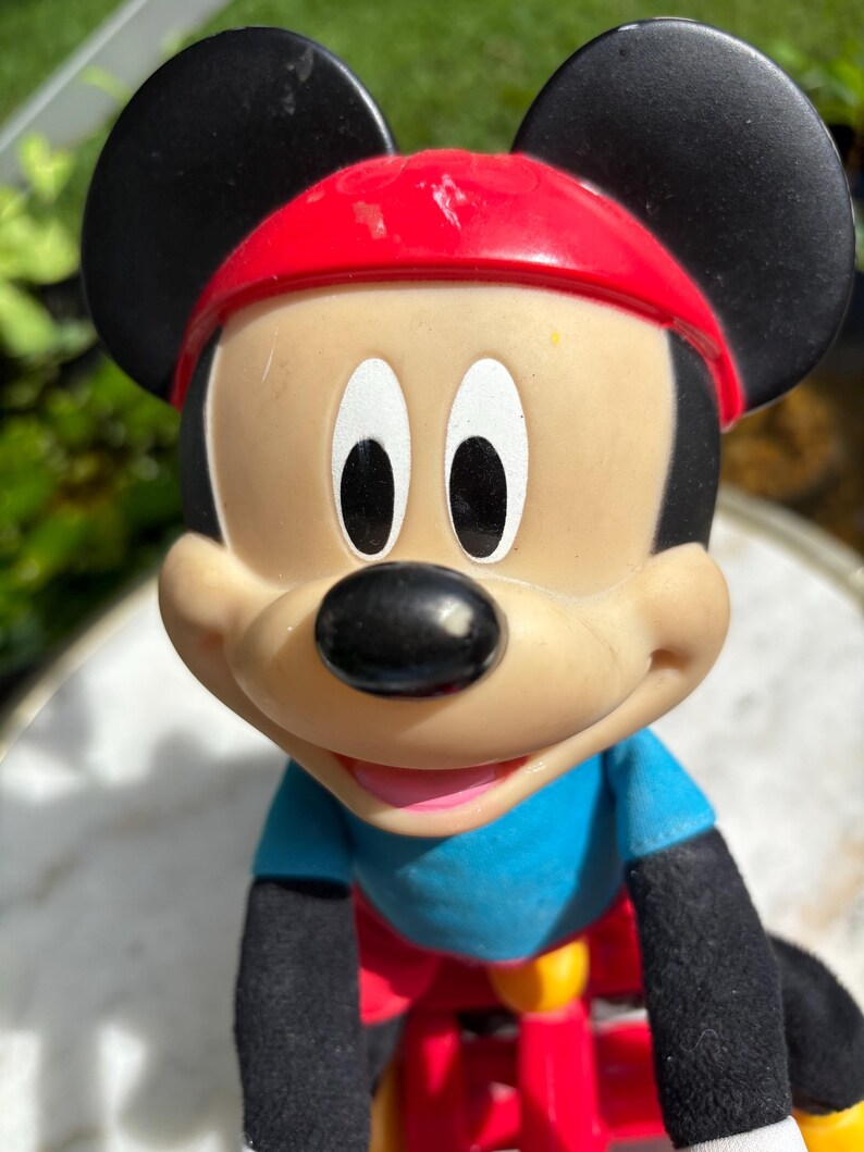 May include: A Mickey Mouse figurine with a red cap, black ears, and a blue shirt. The toy is riding a red and yellow bicycle. The figure has a black nose and white eyes.