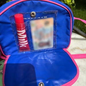 Op de afbeelding: Een blauwe en roze schoudertas met een doorzichtig vakje met een rode lipgloss tube met het label "MILK". De tas heeft een drukknoopsluiting en een roze band. De tekst "LONG THE DOTTED LINE TO OPEN" is zichtbaar.