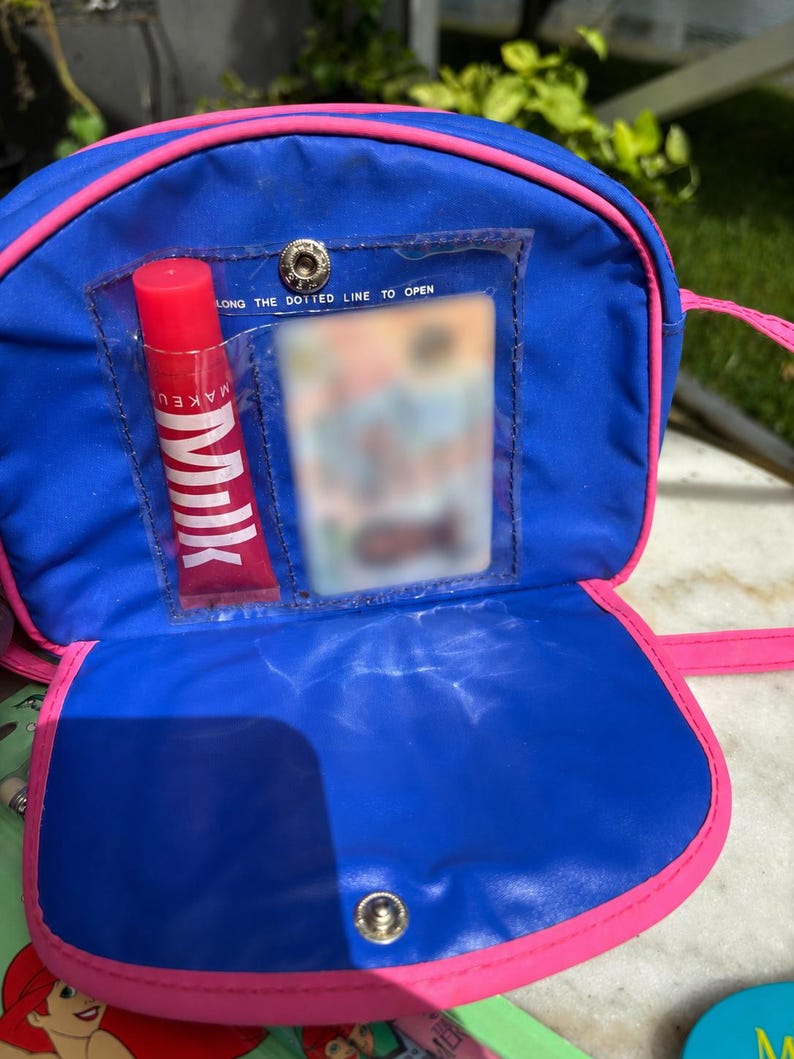 Op de afbeelding: Een blauwe en roze schoudertas met een doorzichtig vakje met een rode lipgloss tube met het label "MILK". De tas heeft een drukknoopsluiting en een roze band. De tekst "LONG THE DOTTED LINE TO OPEN" is zichtbaar.