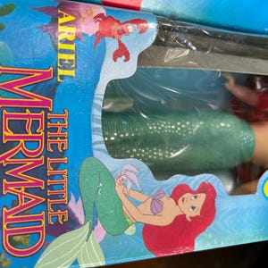 May include: A boxed Little Mermaid toy set. The packaging features Ariel, a red-haired mermaid, and the text "The Little Mermaid." The box is blue with illustrations of Ariel and a crab. The text "Ages 3 and up" is also visible.