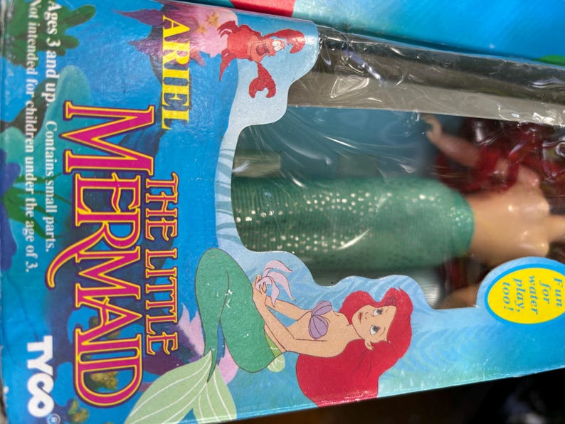 May include: A boxed Little Mermaid toy set. The packaging features Ariel, a red-haired mermaid, and the text "The Little Mermaid." The box is blue with illustrations of Ariel and a crab. The text "Ages 3 and up" is also visible.