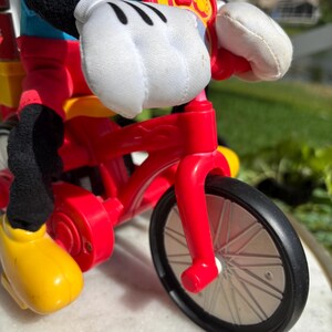 May include: A red toy bicycle with black wheels and a yellow seat. The bicycle is being ridden by a Mickey Mouse plush toy, with white gloves and yellow shoes. The bicycle has a red frame and a black tire.