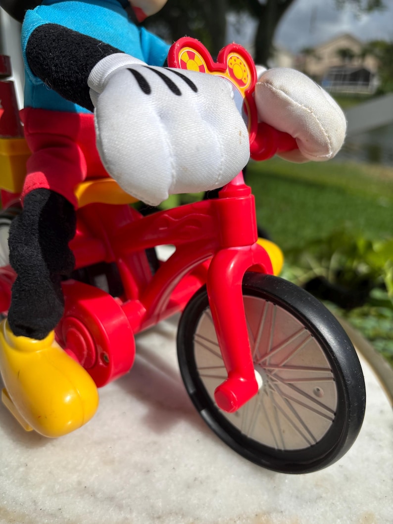 May include: A red toy bicycle with black wheels and a yellow seat. The bicycle is being ridden by a Mickey Mouse plush toy, with white gloves and yellow shoes. The bicycle has a red frame and a black tire.