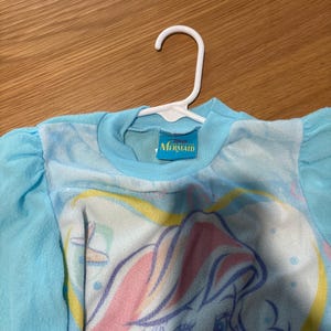 May include: A light blue long-sleeved shirt featuring a graphic of a mermaid with pink and yellow hair. The shirt has a round neckline and is hanging on a white hanger. The tag reads "Mermaid".