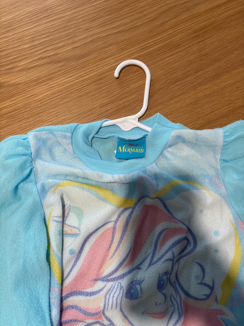 May include: A light blue long-sleeved shirt featuring a graphic of a mermaid with pink and yellow hair. The shirt has a round neckline and is hanging on a white hanger. The tag reads "Mermaid".