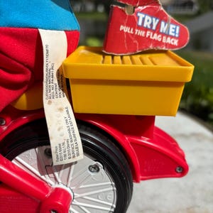 May include: A close-up of a red and yellow toy tricycle with a black tire and white spokes. The tricycle has a yellow storage compartment and a red tag that says "TRY ME! PULL THE FLAG BACK".