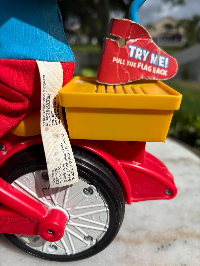 May include: A close-up of a red and yellow toy tricycle with a black tire and white spokes. The tricycle has a yellow storage compartment and a red tag that says "TRY ME! PULL THE FLAG BACK".