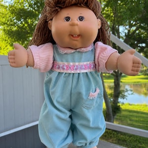 May include: A doll with brown yarn hair, dark eyes, and a single visible tooth. The doll wears a light blue corduroy jumpsuit with a pink butterfly-patterned ribbon at the waist, a pink lace-trimmed collar, and pink short-sleeved undershirt.