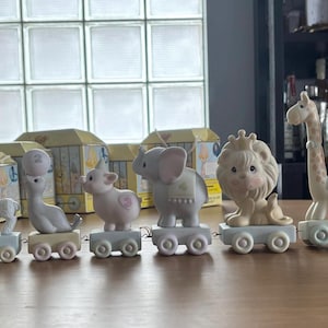 May include: A collection of Precious Moments figurines, including a clown and various animals on train cars. The figurines are pastel-colored and feature a whimsical design. The train cars are light blue and have small wheels. The background includes boxes and a window.
