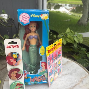 Pode incluir: Uma boneca Ariel de A Pequena Sereia da Disney da Tyco numa caixa azul, um cart&atilde;o vertical com tr&ecirc;s bot&otilde;es circulares da Ariel e uma folha de autocolantes Disney Star com v&aacute;rias ilustra&ccedil;&otilde;es da Ariel e do Linguado do filme.