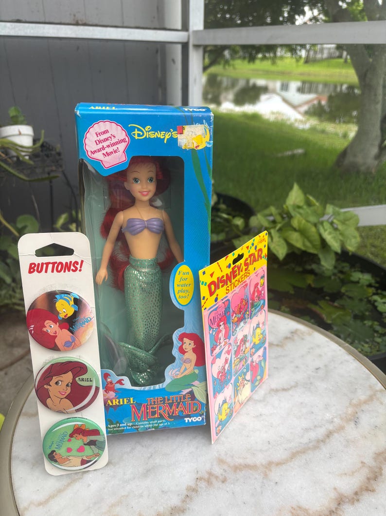 Pode incluir: Uma boneca Ariel de A Pequena Sereia da Disney da Tyco numa caixa azul, um cart&atilde;o vertical com tr&ecirc;s bot&otilde;es circulares da Ariel e uma folha de autocolantes Disney Star com v&aacute;rias ilustra&ccedil;&otilde;es da Ariel e do Linguado do filme.