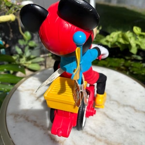 May include: A Mickey Mouse toy figure riding a red and yellow tricycle. The figure has black ears, a red head, blue shirt, and black pants. The tricycle has white tires and yellow foot rests. A blue and yellow flag is attached to the back.