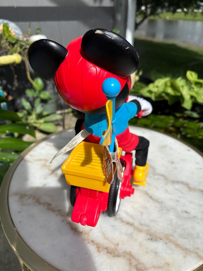 May include: A Mickey Mouse toy figure riding a red and yellow tricycle. The figure has black ears, a red head, blue shirt, and black pants. The tricycle has white tires and yellow foot rests. A blue and yellow flag is attached to the back.