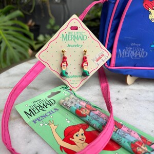 Op de afbeelding: Een verzameling vintage "De Kleine Zeemeermin" merchandise, waaronder oorbellen, potloden en een blauwe rugzak. De oorbellen tonen Ariel, de zeemeermin, met rood haar en een groene staart. De potloden zijn verpakt in een groene kaart met Ariel-illustraties.
