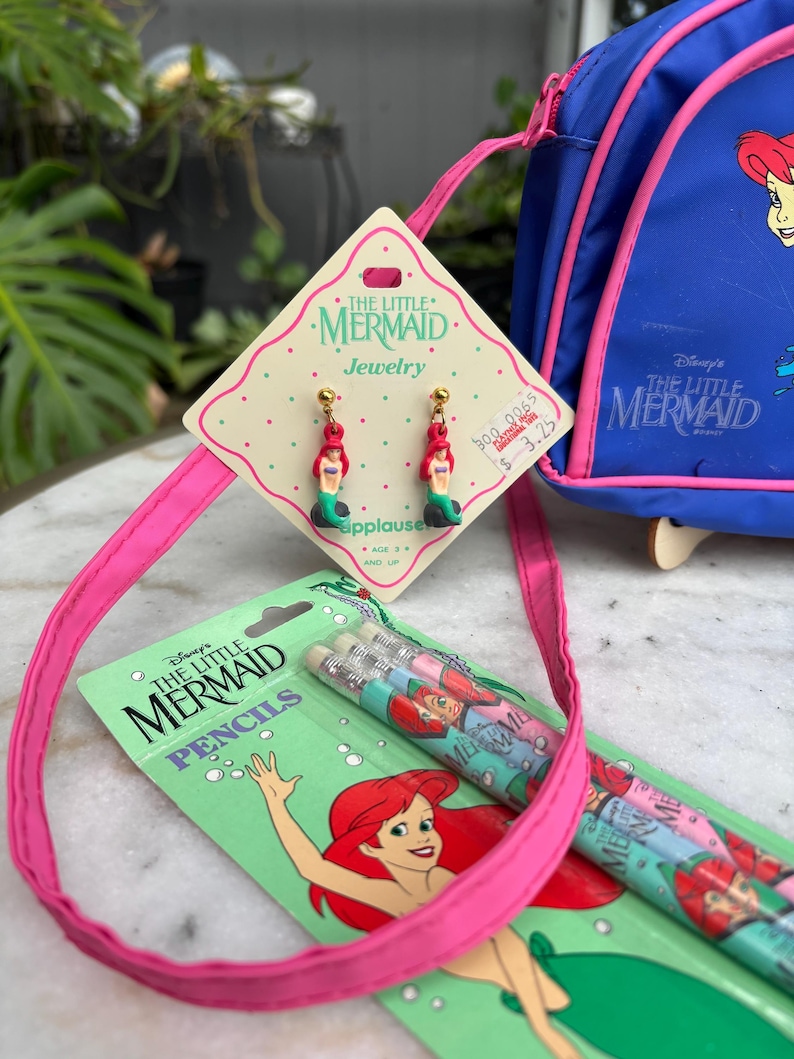 Op de afbeelding: Een verzameling vintage "De Kleine Zeemeermin" merchandise, waaronder oorbellen, potloden en een blauwe rugzak. De oorbellen tonen Ariel, de zeemeermin, met rood haar en een groene staart. De potloden zijn verpakt in een groene kaart met Ariel-illustraties.