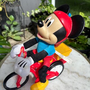 May include: A Mickey Mouse figurine riding a red tricycle with a yellow cargo box. Mickey wears a red helmet, blue shirt, black gloves, red pants, and yellow shoes. The tricycle has white tires and a white handlebar.