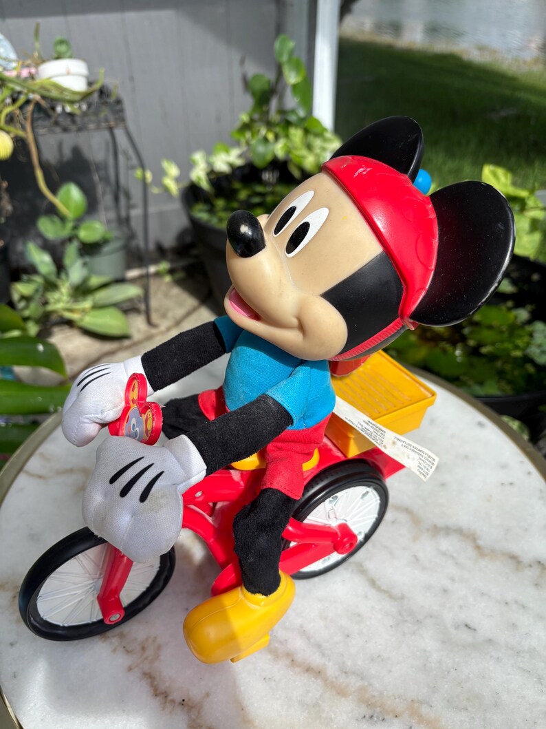 May include: A Mickey Mouse figurine riding a red tricycle with a yellow cargo box. Mickey wears a red helmet, blue shirt, black gloves, red pants, and yellow shoes. The tricycle has white tires and a white handlebar.