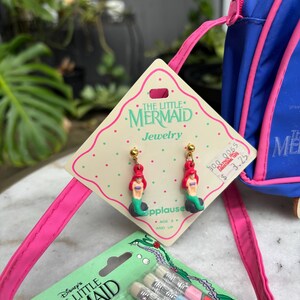 Op de afbeelding: De afbeelding toont vintage sieraden en potloden met het thema "De Kleine Zeemeermin". De oorbellen hebben Ariel-figuren met rood haar en groene staarten. De potloden zitten in een groene verpakking met een afbeelding van Ariel.