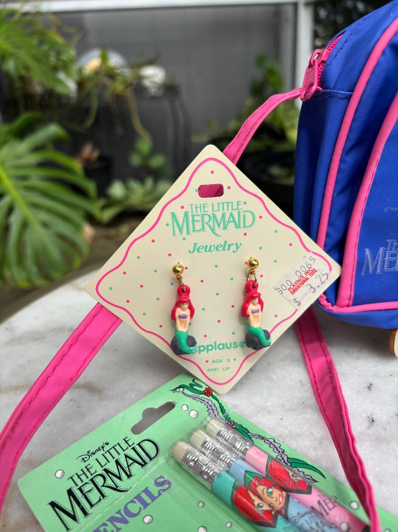 Op de afbeelding: De afbeelding toont vintage sieraden en potloden met het thema "De Kleine Zeemeermin". De oorbellen hebben Ariel-figuren met rood haar en groene staarten. De potloden zitten in een groene verpakking met een afbeelding van Ariel.